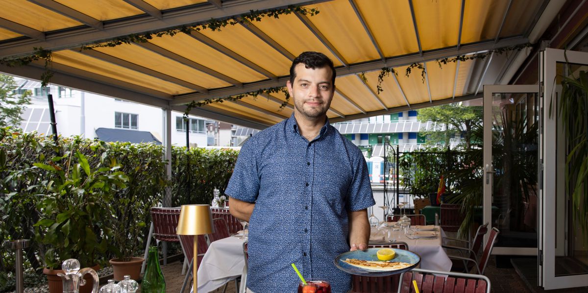 Ein Kellner in einem Restaurant serviert lächelnd zwei Teller mit Gerichten unter einer überdachten Terrasse.