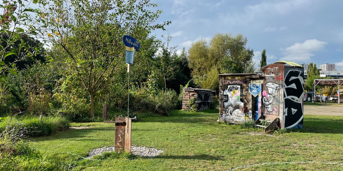 Ein gepflegter Garten mit Hochbeeten und Sonnenblumen im Vordergrund. Im hinteren Bereich des Gartens steht ein Tisch mit Stühlen unter einem kleinen Pavillon. Die Umgebung ist grün und von Bäumen umgeben.