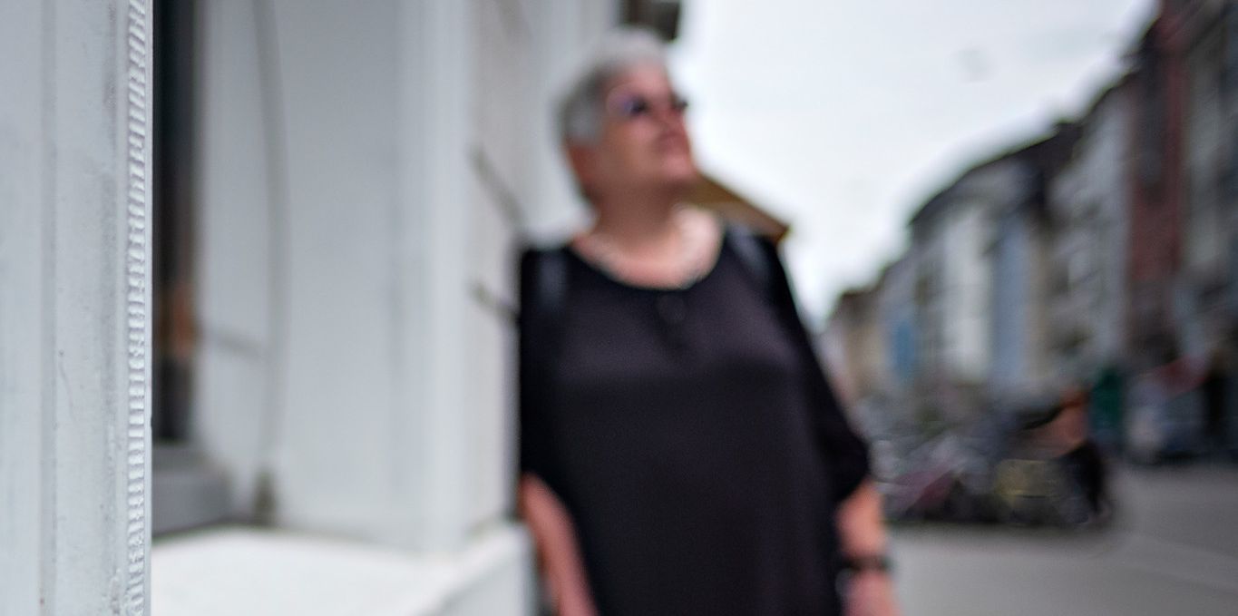 Eine Frau mit weissem Haar lehnt sich an die Wand eines Gebäudes in einer unscharfen Strasse. Im Hintergrund sind Gebäude und der Himmel sichtbar.