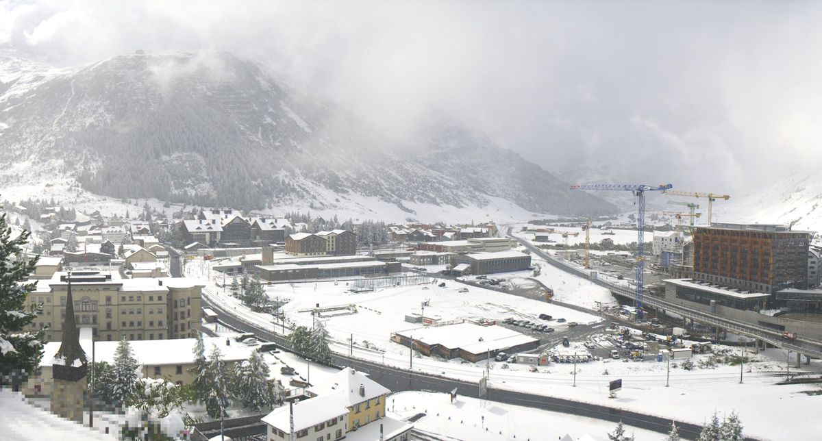 Schnee bedeckt Andermatt am 13. September um 12:40 Uhr. Der Ort zeigt verschneite Gebäude, Strassen und Bäume, während im Hintergrund Baukräne und ein bewölkter Himmel sichtbar sind.