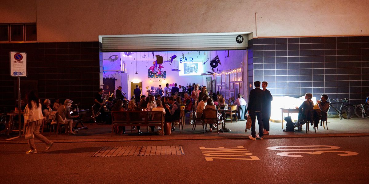 Menschen sitzen an Tischen vor einer belebten Bar in einer urbanen Strasse bei Nacht. Die Bar ist durch bunte Beleuchtung und Wanddekorationen belebt.