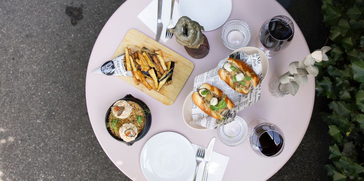 Essensarrangement aus der Vogelperspektive mit zwei belegten Sandwiches, Pommes frites auf einem Holzbrett und einem Salat auf einem runden Tisch im Freien