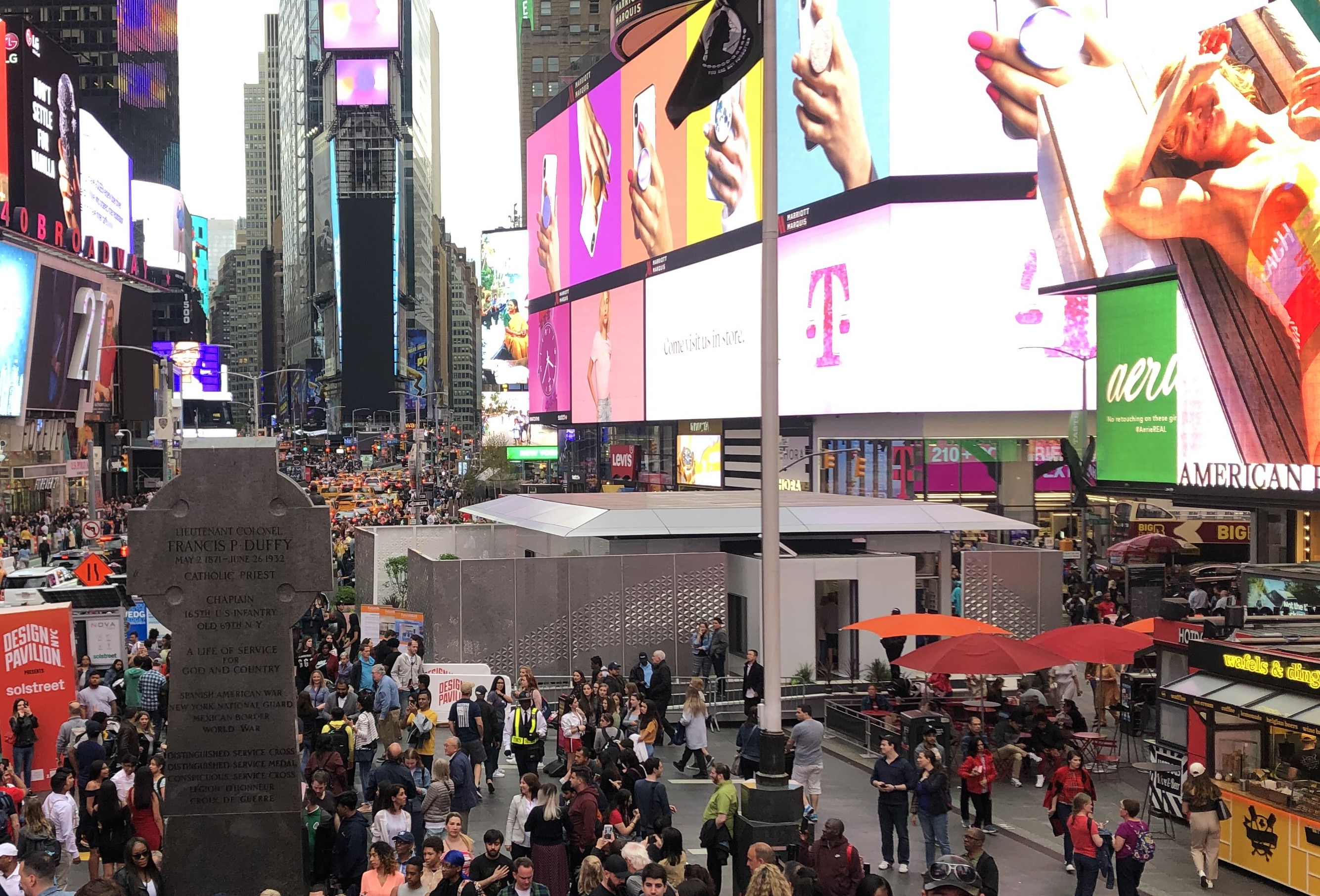 The pedestrian plaza in Times Square is full of people who are gathered around a solar powered prefab home. The billboards on the surrounding buildings are all brightly lit.