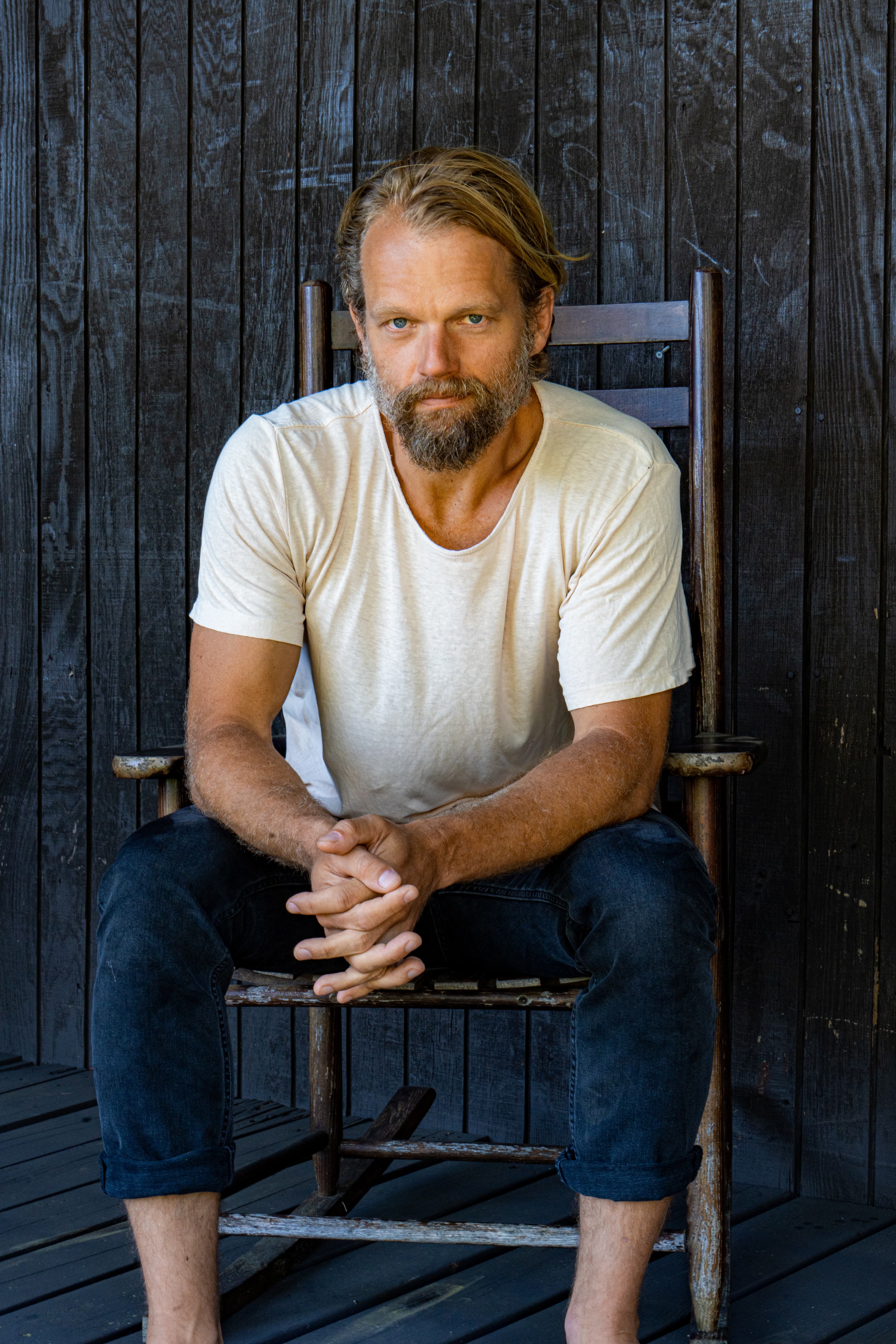 A portrait of Travis Lett wearing a white t-shirt with a beard and longish hair.