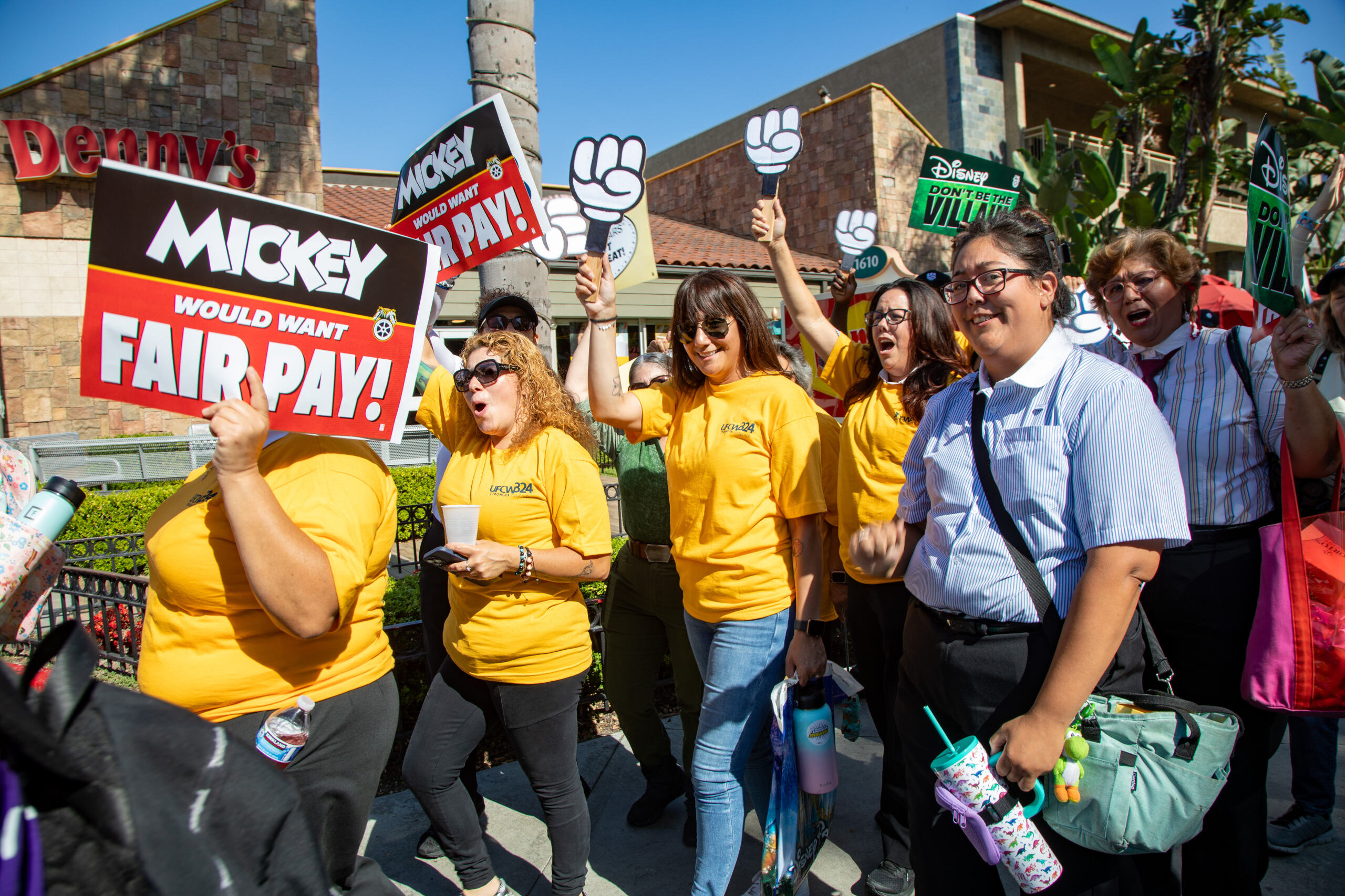 Workers in yellow t-shirts protest outside Disneyland with mouse mittens and signs that say Mickey would want fair pay