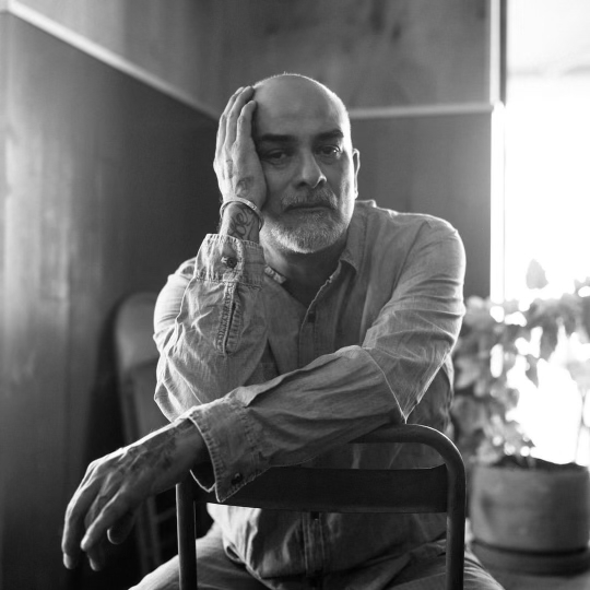 Steven Arroyo in black and white holding his hand to his temple in a chair