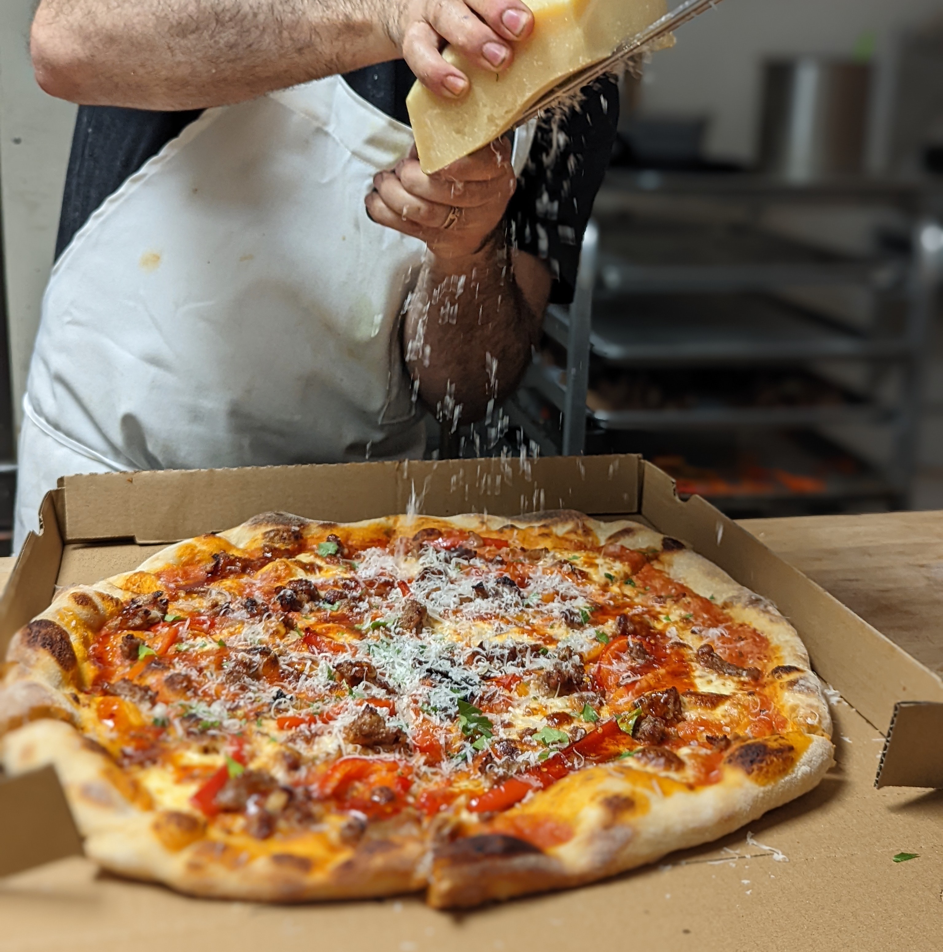 A hand shaves down pieces of dry cheese on top of a boxed pizza.