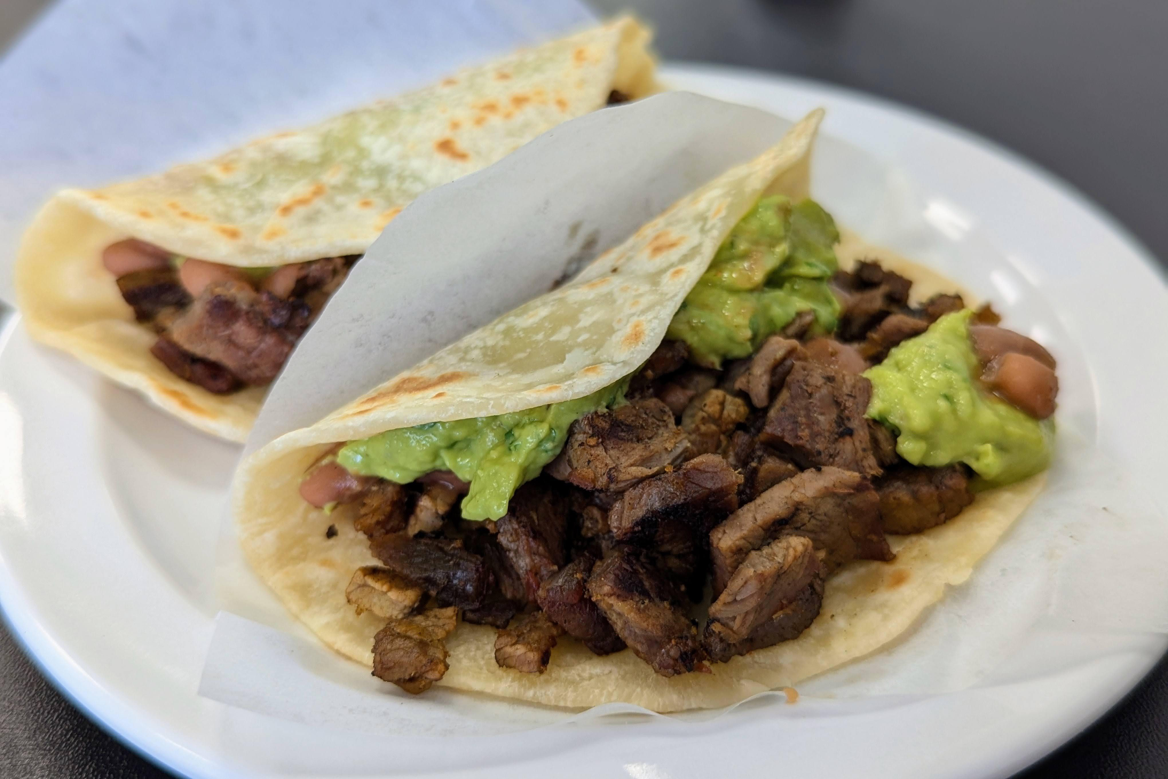 Two asada tacos on tortillas wrapped in white paper on a white plate from Asadero Chikali