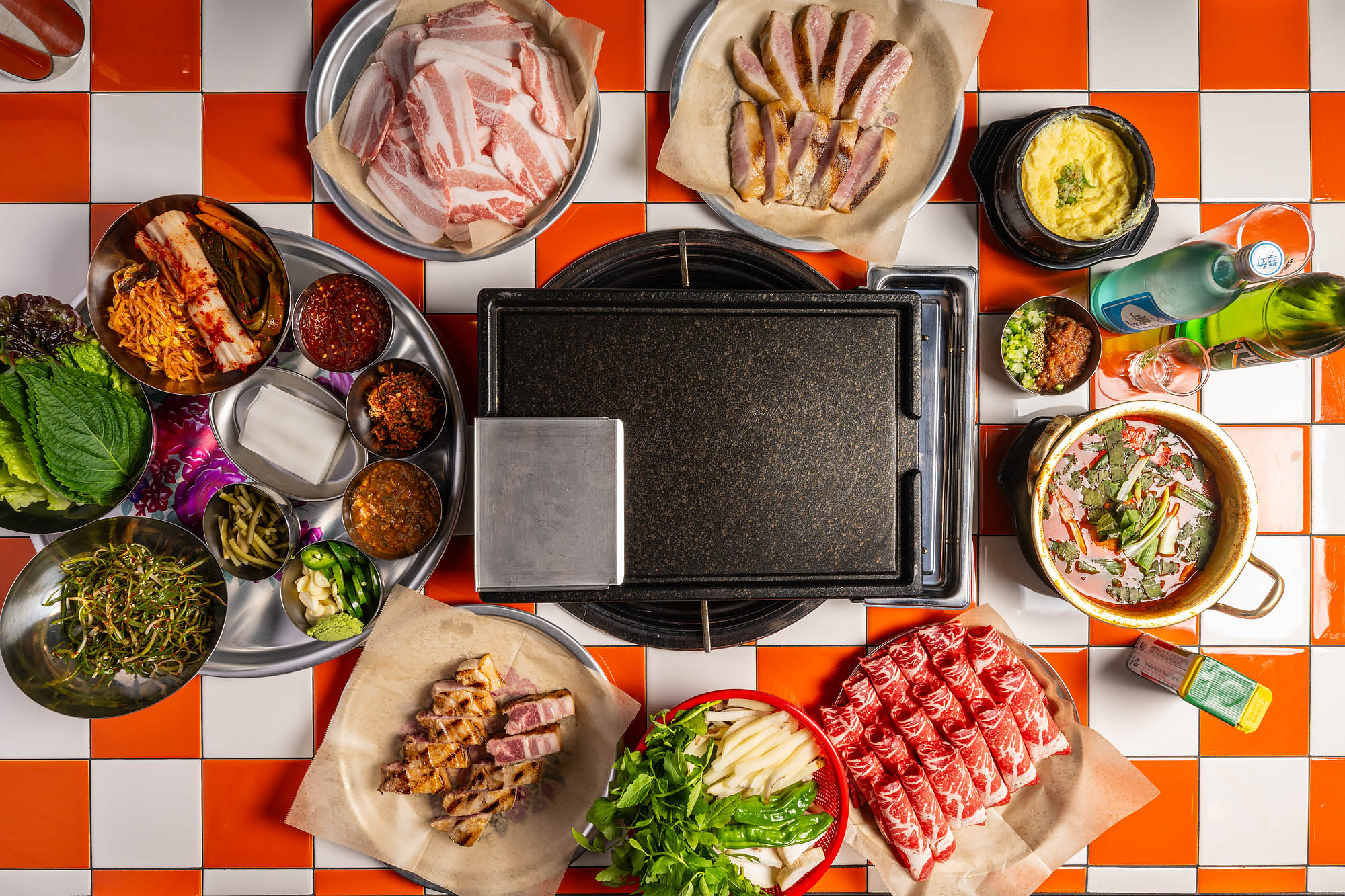 Uncooked meats, side dishes, and soju with a cast iron grill in the center.