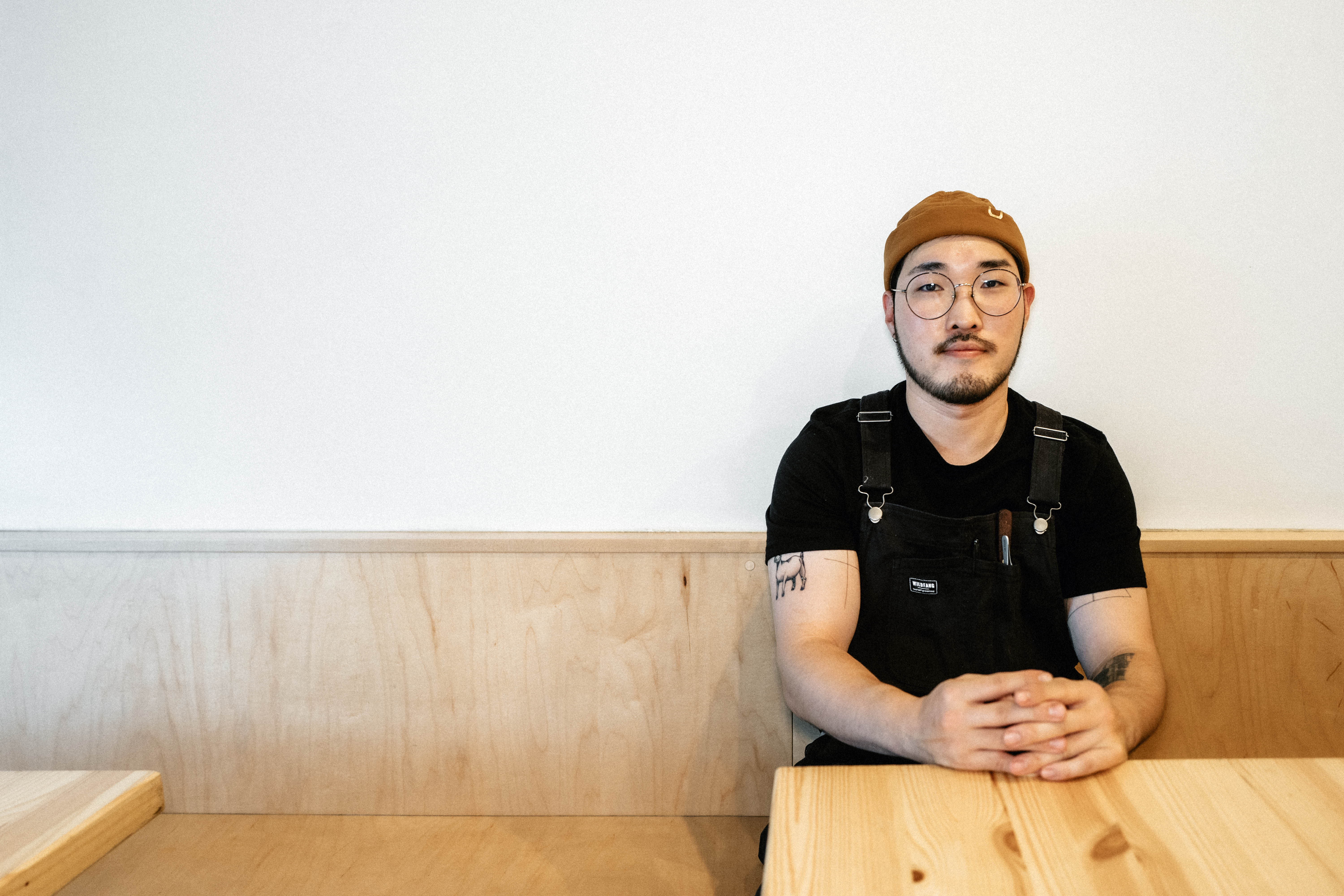 A male Korean American chef wearing a brown beanie sits with hands folded at a restaurant table.