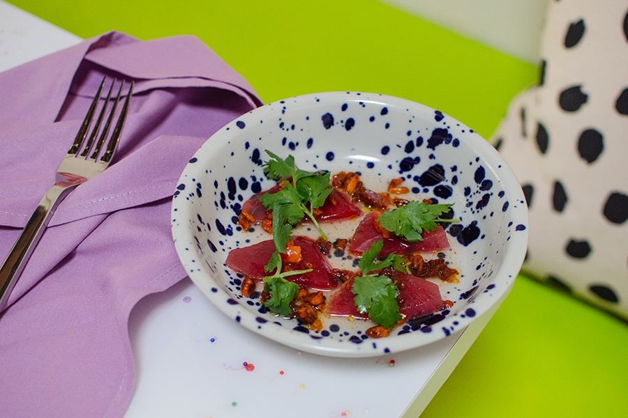 Four slices of raw tuna with herbs and condiments sit in a white bowl with blue dots on a white table with colorful dots and a lavender napkin.