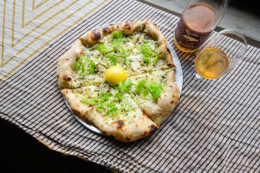 A white pizza with greens and a lemon wedge sits on a striped cloth next to a glass and bottle of wine.