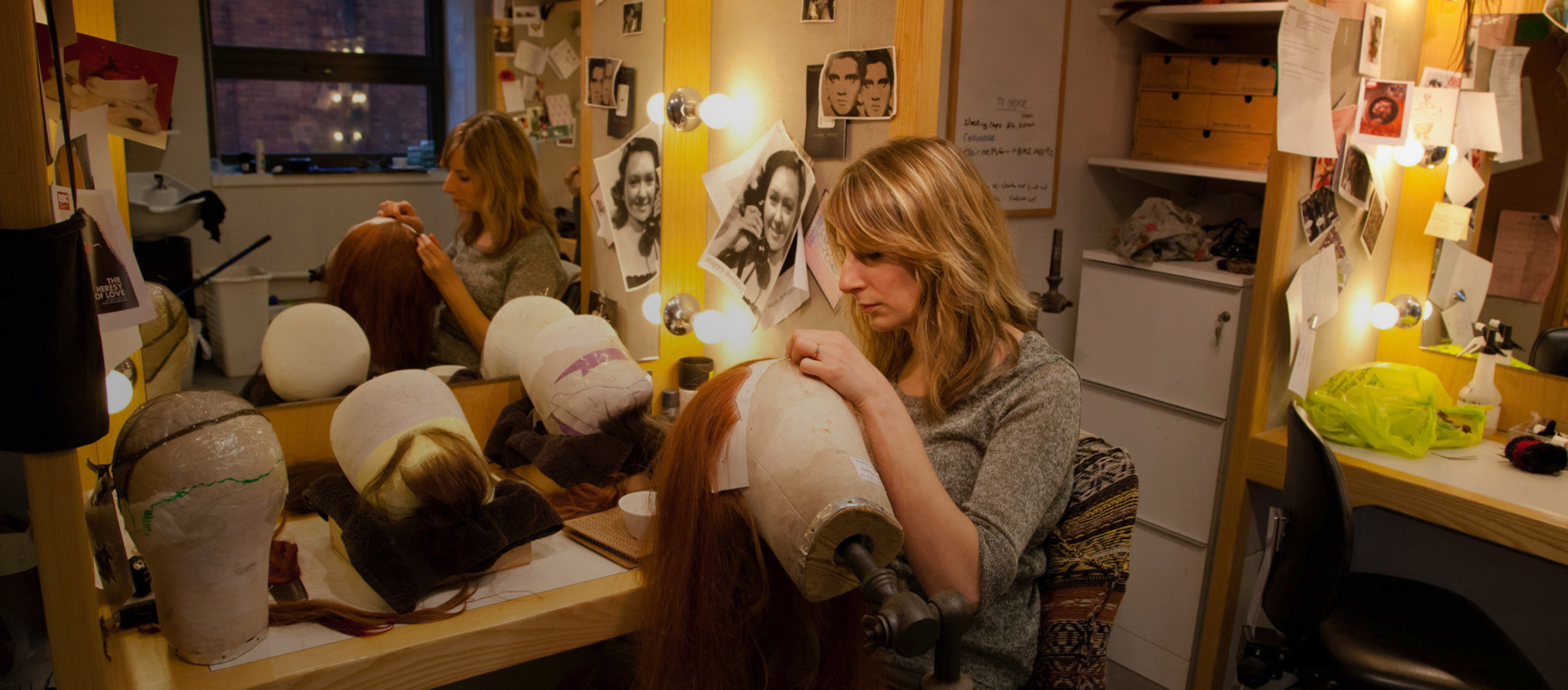 A blonde woman holding a head model and stitching a long red wig in a dressing room full of photos and wigs