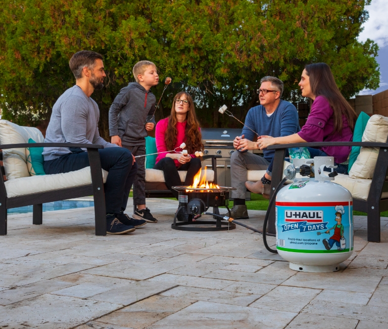 A family sits around a propane fire roasting marshmallows on skewers, watching their son blow the flame off his marshmallow