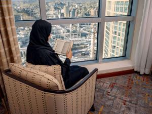 uma mulher sentada numa cadeira a ler um livro numa janela em Swissotel Al Maqam Makkah em Meca