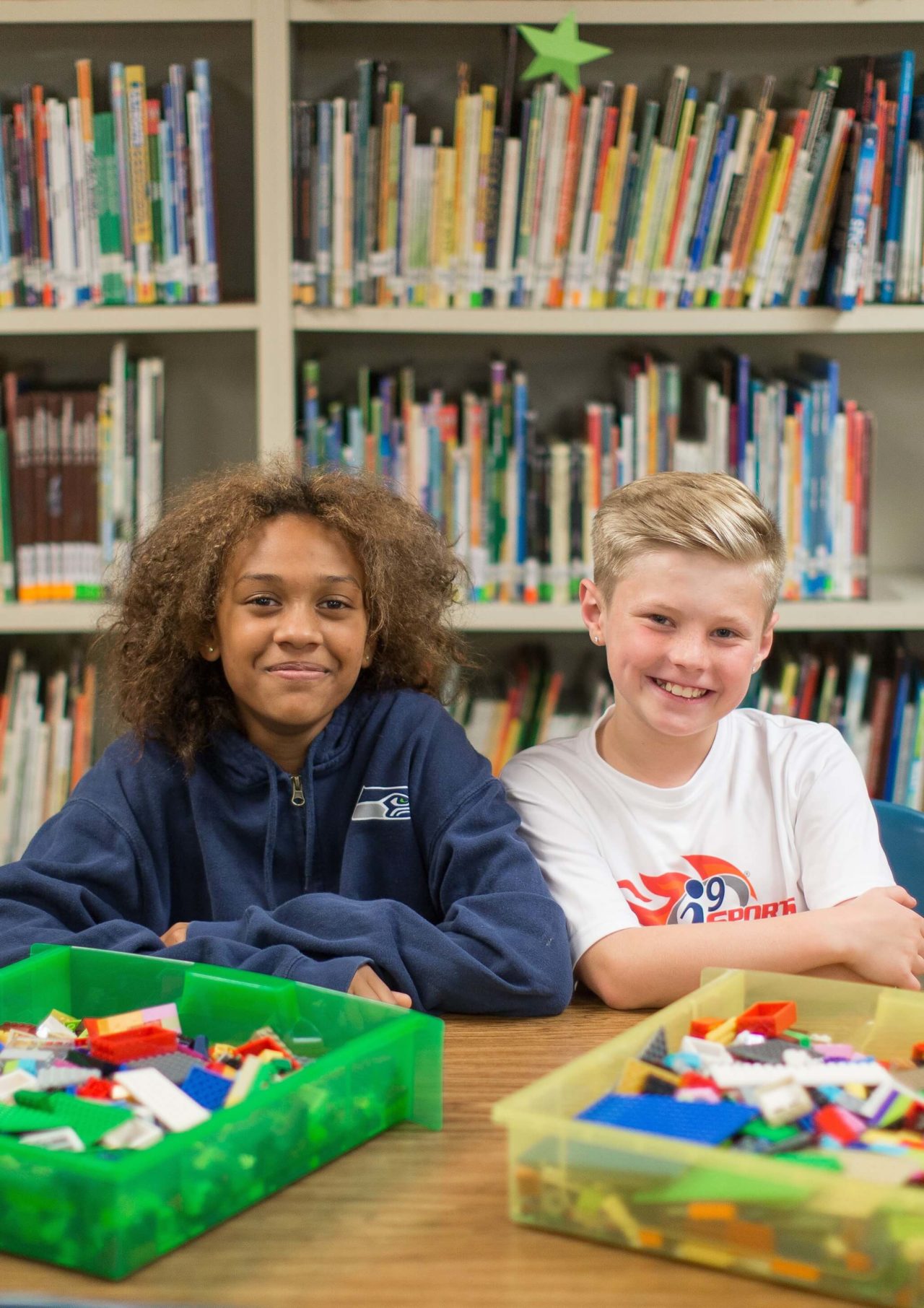 Two smiling students sitting in front of a bookshelf