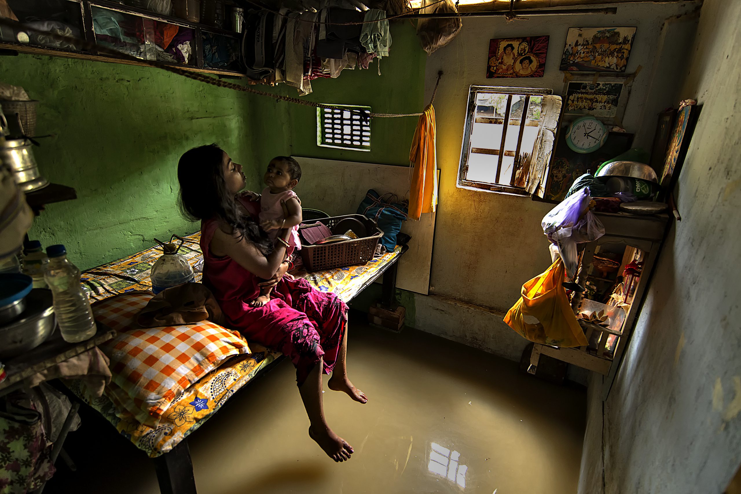 A mother and baby sitting on a bed above floodwater in their flooded home.