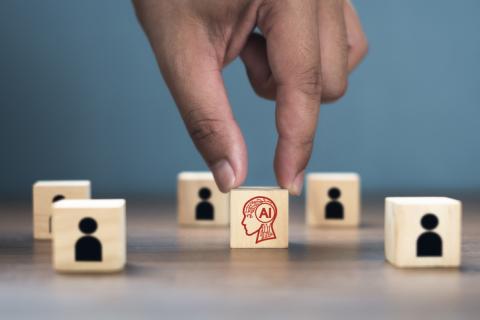 A hand reaches for a wooden block displaying "AI"