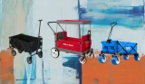 A black, red and blue collapsible folding wagon stand side by side on a blue, orange and white painted background