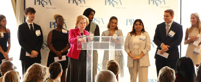 Dana Clay, in red, daughter of Ev Clay, addresses the audience while Diana La Torre, Sebastian Gonzalez, and Adelaide Doman stand in the back toward the right during the 37th Annual Ev Clay/PRSA Miami Chapter Endowment Fund Luncheon held at the Rusty Pelican on Oct. 4.