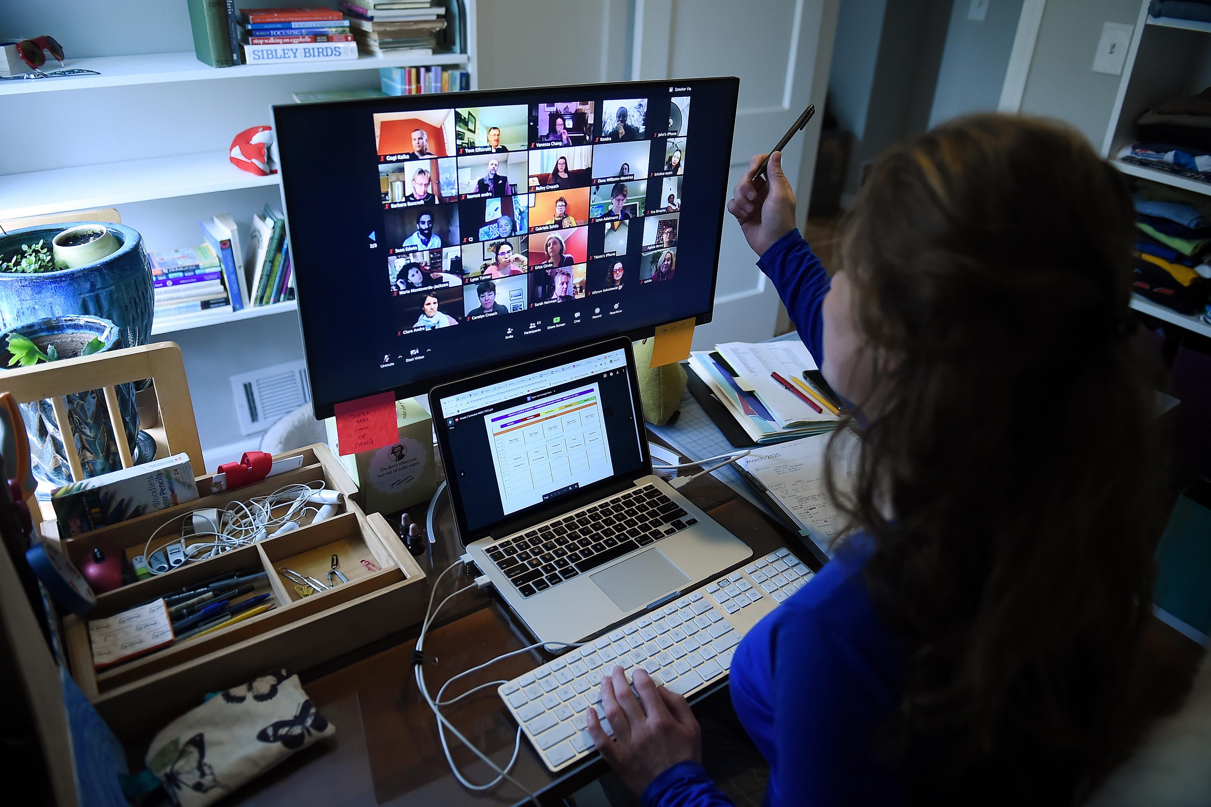 A woman looks at a screen—the back of her head is visible. She has her laptop before her and a monitor featuring Zoom, with windows of people staring back at her.