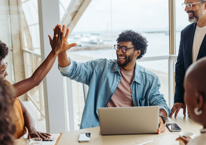 Cinco colegas de trabalho reunidos. Um deles está comemorando a retrospectiva de 2024 do Mercado Pago, dando um high-five em outro colega, enquanto está na frente do computador.