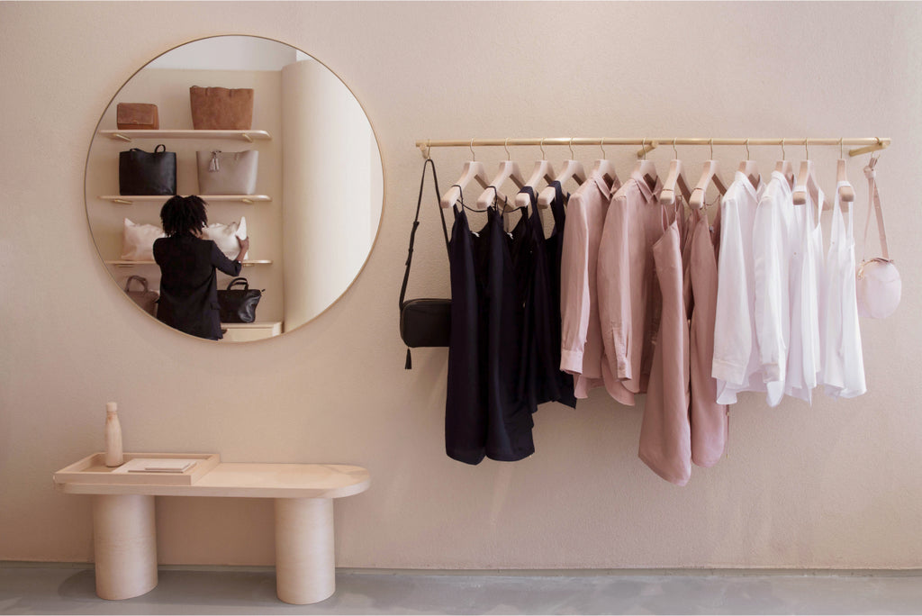 A clothing store interior with dresses on hangers, a bench, and a woman reflected in a round mirror.