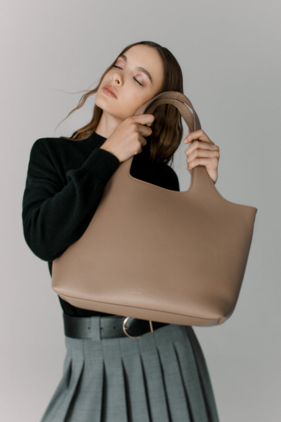 Person holding a large handbag over shoulder, eyes closed, wearing a sweater and pleated skirt.