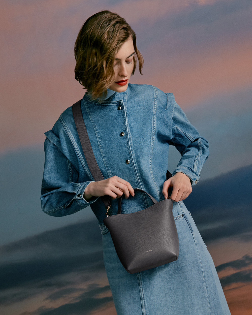 Woman in a denim outfit holding a handbag, standing against a sky background.