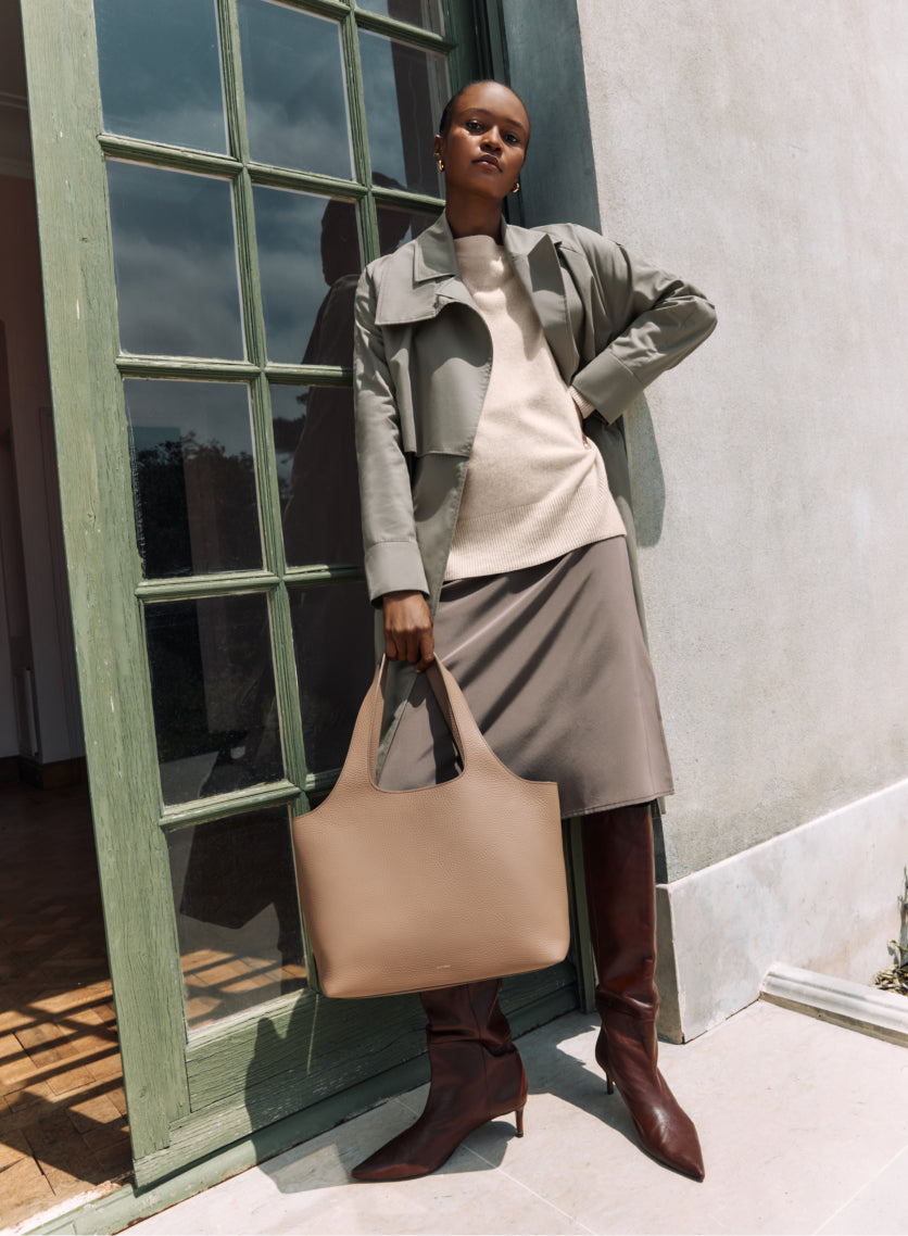 Person standing by a window with a bag and wearing a jacket, sweater, skirt, and boots.