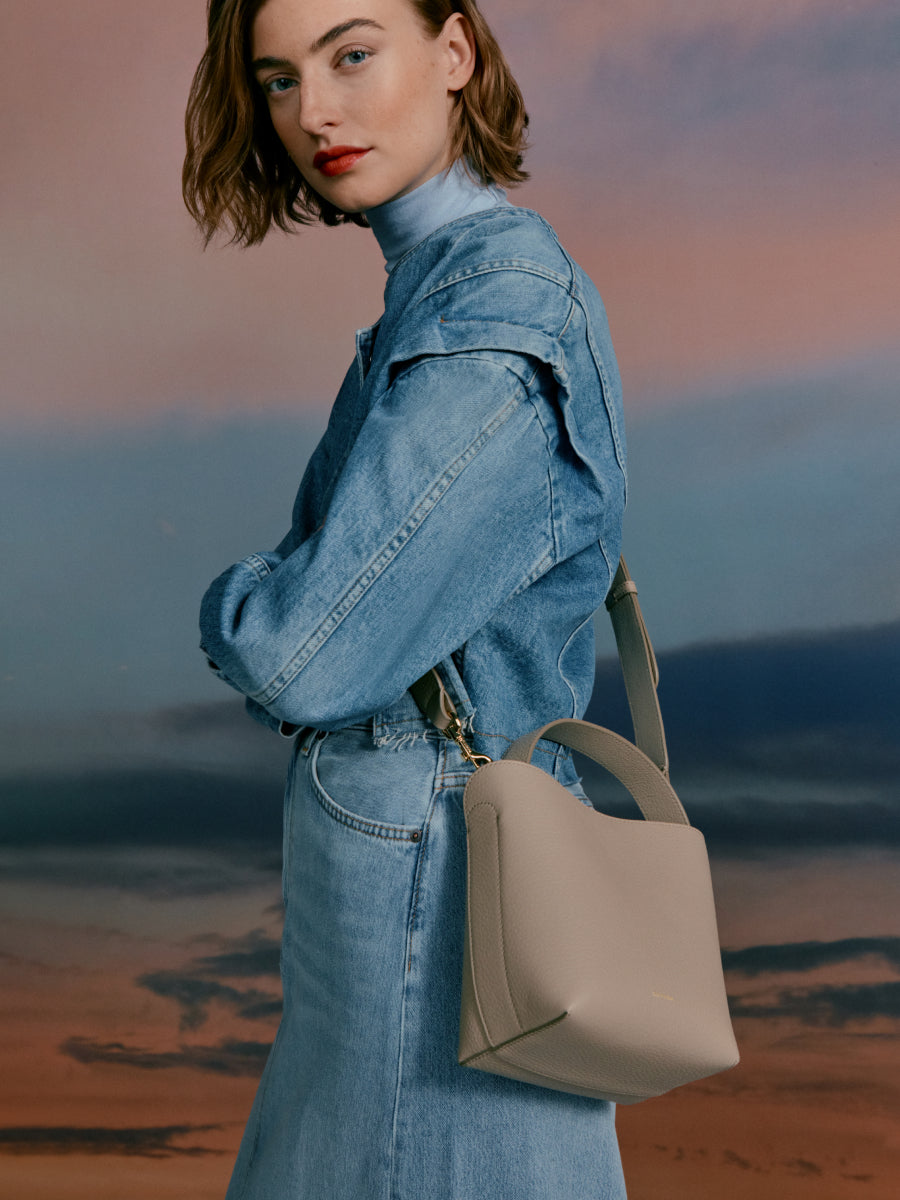 Woman wearing a denim outfit carrying a handbag, posing against a sunset backdrop.