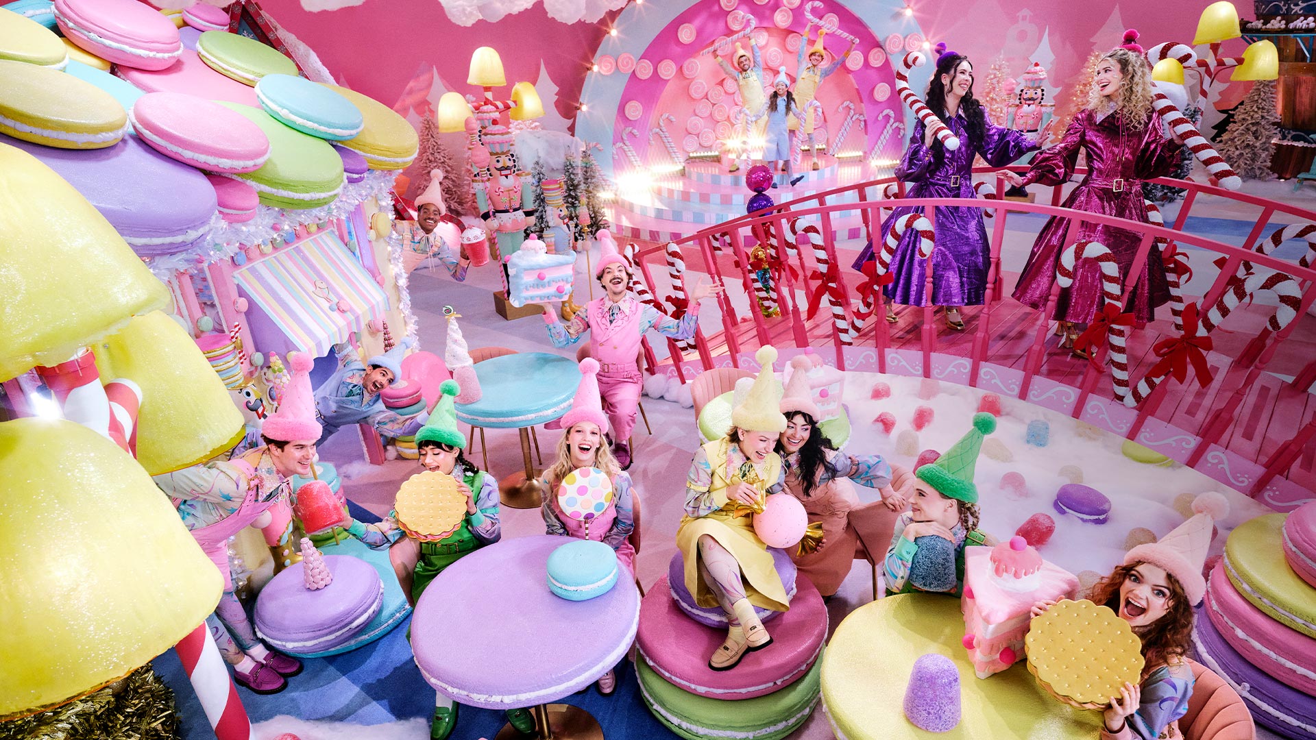 A colorful candy-themed scene with people dressed as elves and characters, surrounded by giant sweets like macarons and candy canes, creating a playful, whimsical atmosphere