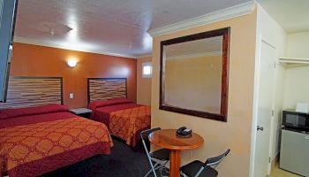 The image shows a hotel room with two double beds, a wall-mounted mirror, a small round table with two chairs, a microwave, and open closet space.