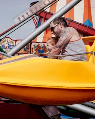 A man and child are riding a yellow amusement park ride, with colorful attractions and structures visible in the background.