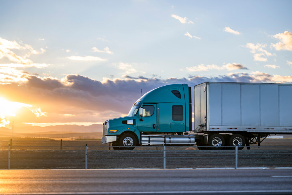 Industrial long hauler big rig green semi truck tractor with chrome parts transporting commercial cargo in loaded dry van semi trailer running on the highway road at sunset time in California