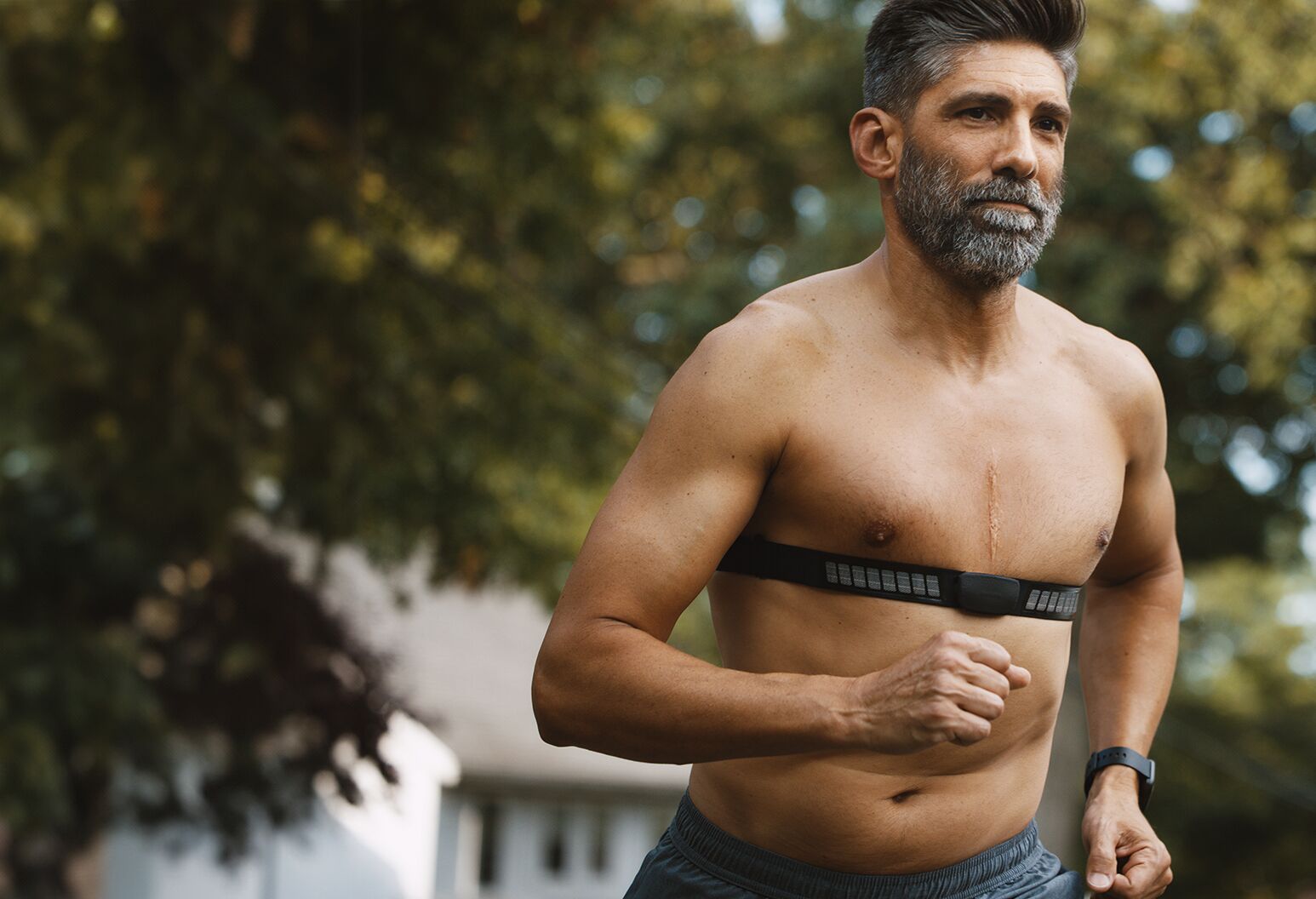 Fit, shirtless man with brown and gray hair jogging with trees in background