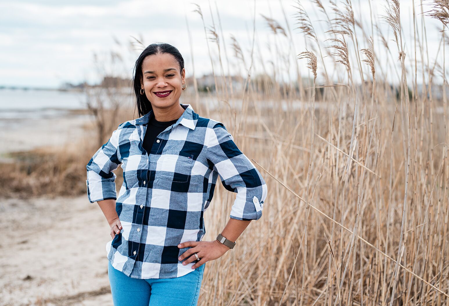 Woman with dark hair, plaid shirt and jeans outside in marshy area.