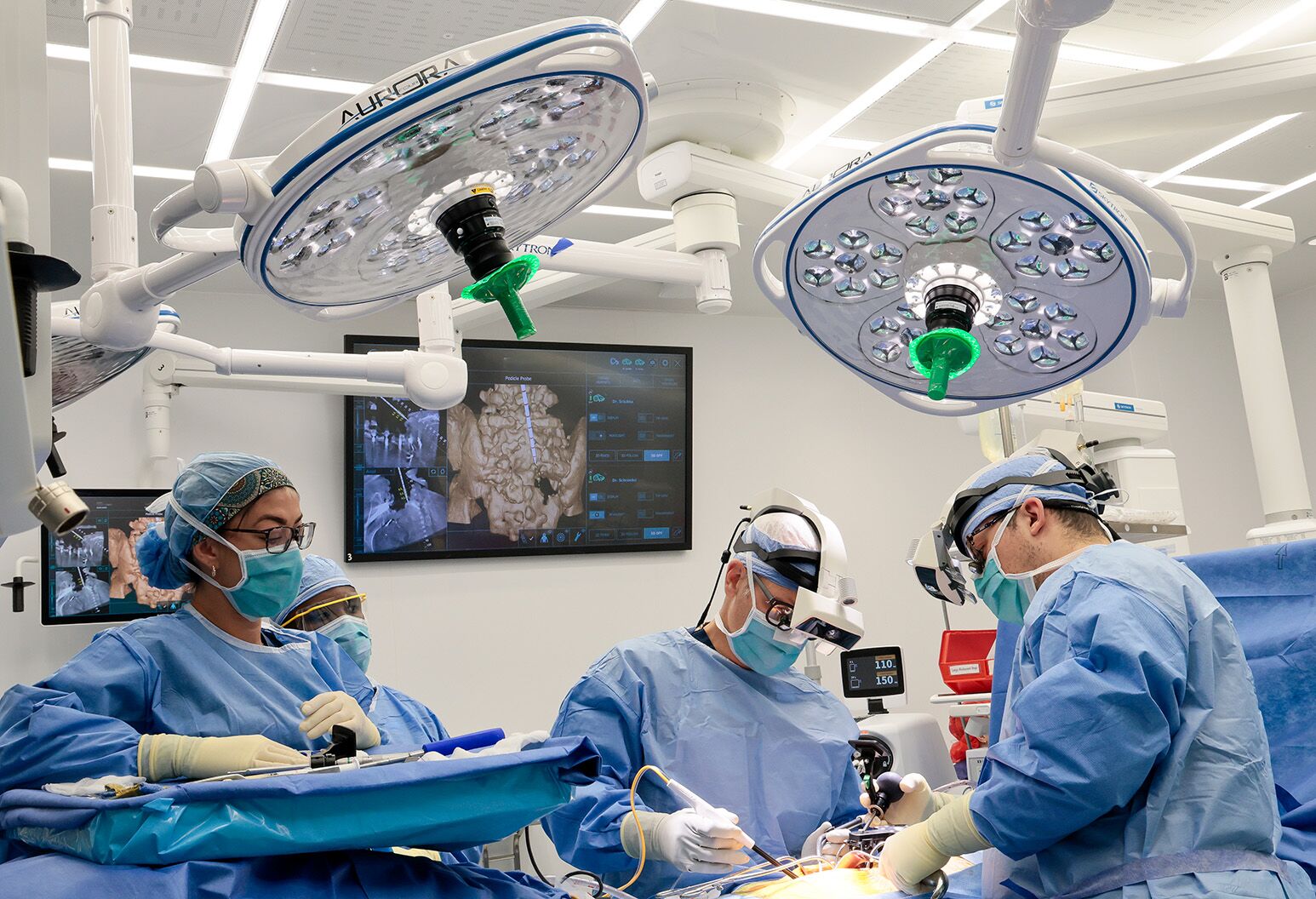 Two surgeons in scrubs and headlamps perform a surgery at right; two more at left stand and observe.