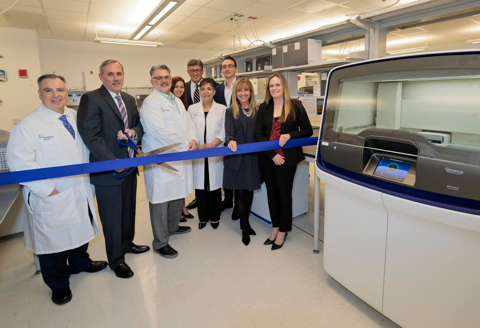 A group of men and women, some in white coats, stands in a medical laboratory for a ceremonial ribbon-cutting.