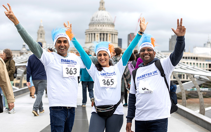 Three people stand on a bridge smiling at the camera with their arms in the air. They're wearing white Diabetes UK t-shirts and blue and orange Diabetes UK hats.