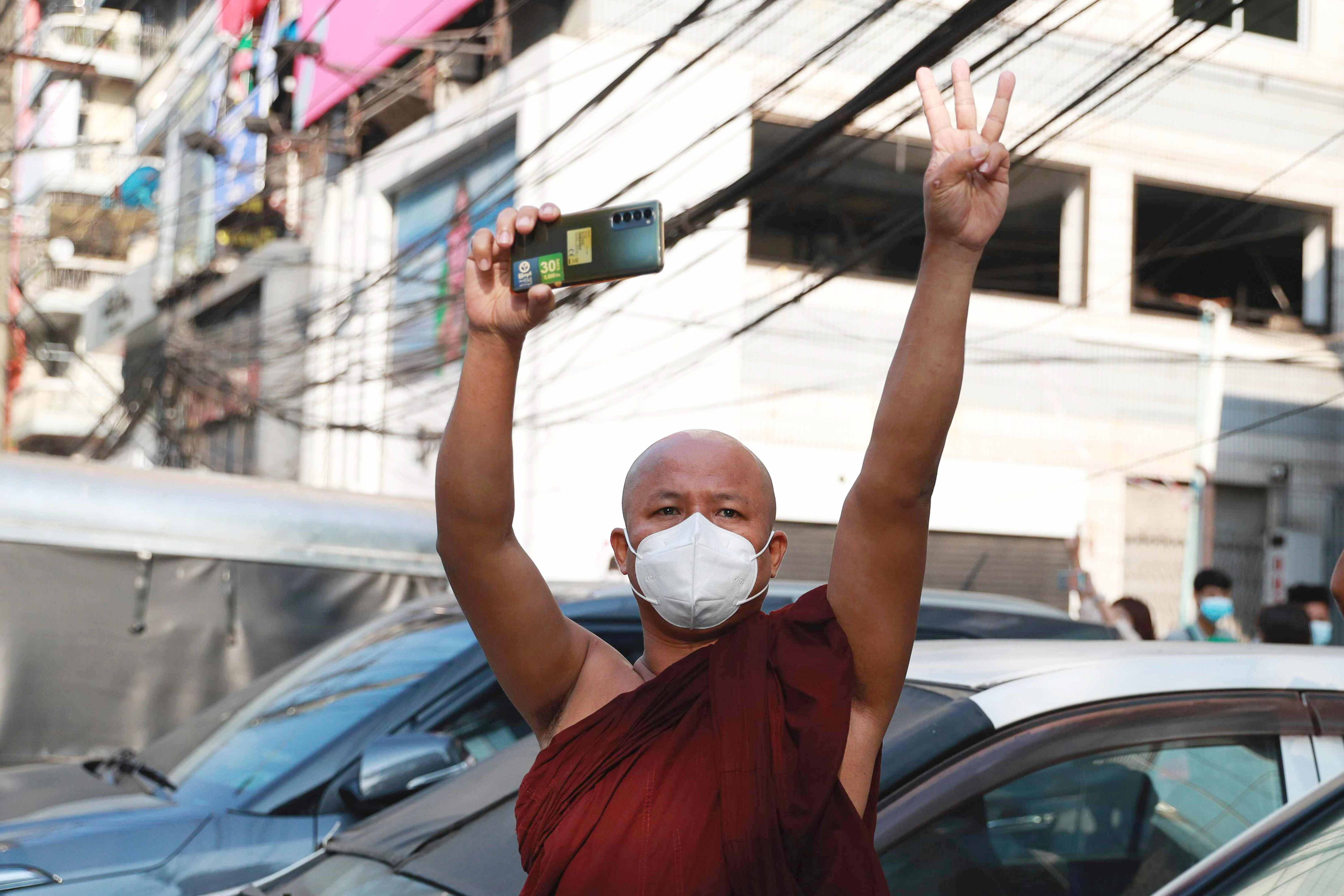 FILE - A Buddhist monk flashes the three-fingered salute holding is smart phone as he watches protesters march in Yangon, Myanmar on Sunday, Feb. 7, 2021. Myanmar’s military government has launched a major effort in June of 2024, to block free communication on the Internet, shutting off access to virtual private networks -- VPNs -- which can be used to circumvent blockages of banned websites and services. (AP Photo, File)