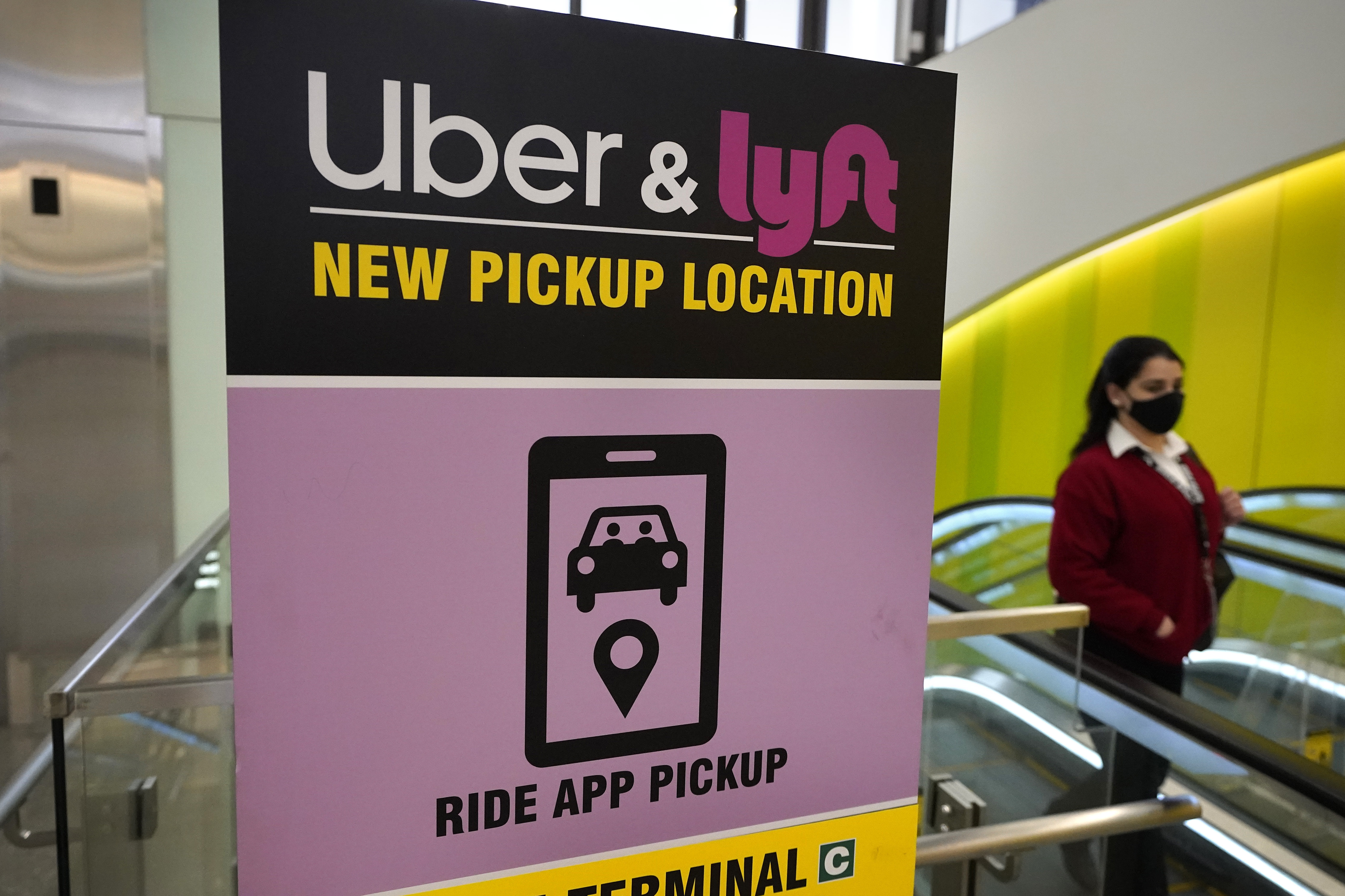 FILE - A passerby walks past a sign offering directions to an Uber and Lyft ride pickup location at an airport, Feb. 9, 2021, in Boston. Drivers for Uber and Lyft will earn a minimum pay standard of $32.50 per hour under a settlement announced, Thursday, June 27, 2024, by Massachusetts Attorney General Andrea Campbell, a deal that also includes a suite of benefits and protections. (AP Photo/Steven Senne, File)