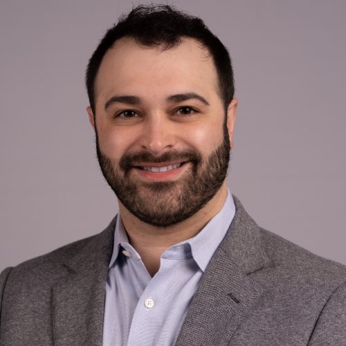 Gabriel Rubinstein is a White man with short black hair and a full beard and mustache. He smiles wearing a grey blazer and light blue collared shirt.