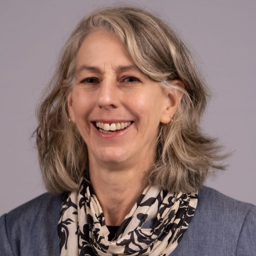 Leslie Dickinson is a White woman with blond-brown long wavy hair. She smiles brightly wearing a blue jacket and black and cream patterned scarf around her neck.