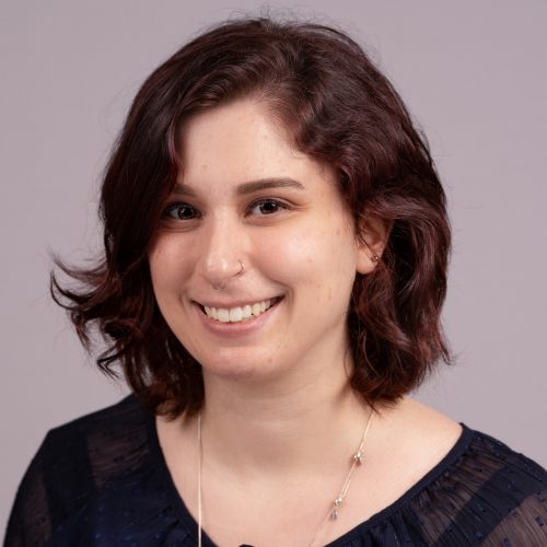 Sara Miller is a young White woman with brunette, shoulder length wavy hair and 2 thin nose rings. She smiles wearing a black and blue blouse.