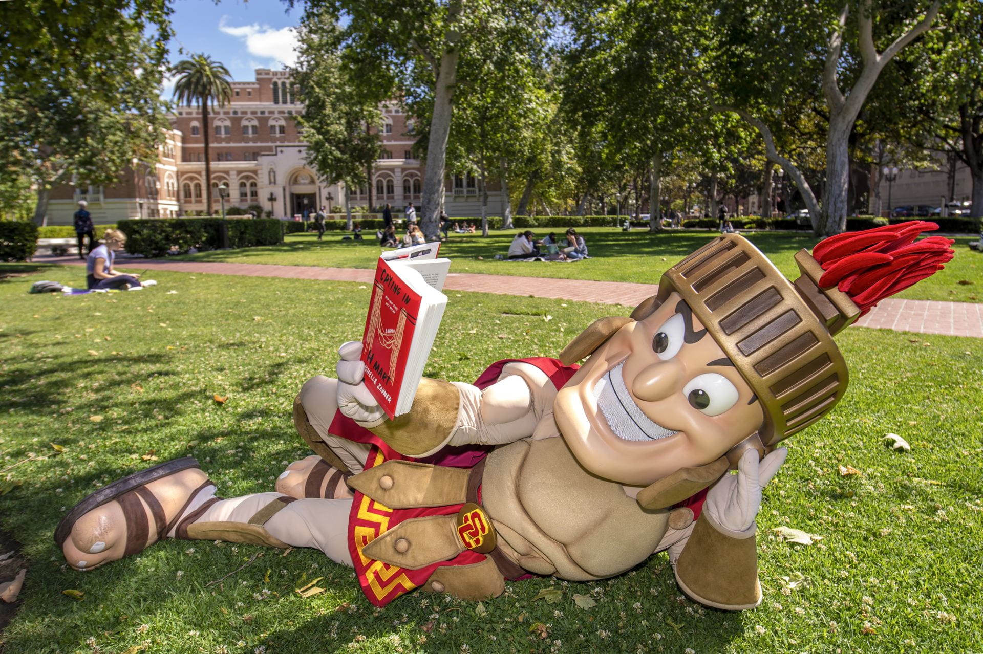 Tommy Trojan mascot reading and relaxing on the grass in Alumni Park.