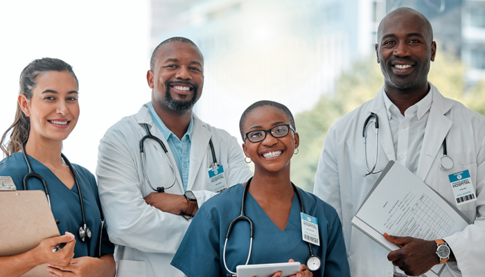 2 women and 2 men in doctor or nurse uniforms smiling at the camera, some holding charges or clip boards, wearing stethoscopes around their necks