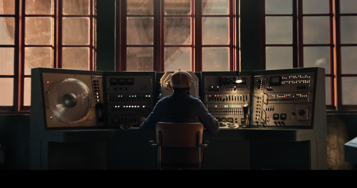 Character sitting in a dark room on a chair looking at a large switchboard.