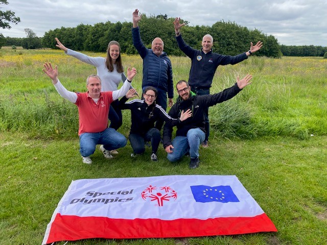 A group of six people stand in a green field with their hands in the air in celebration in front of a Special Olympics flag with the European Union logo.  