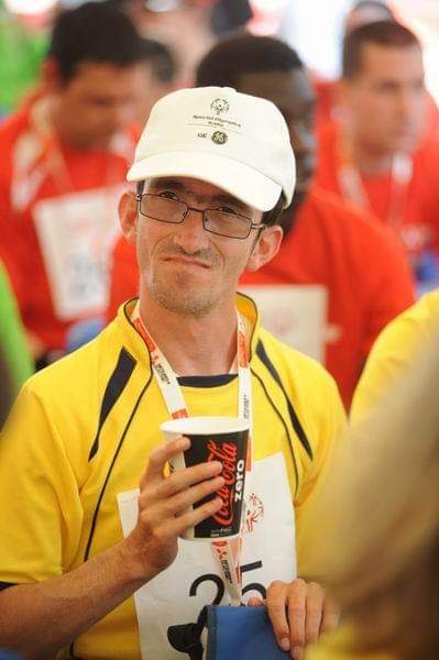 A man in a yellow t-shirt and baseball cap drinks from a Coca Cola branded paper cup.